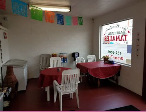 Cozy dining area with red tablecloths, white chairs, and colorful papel picado decorations. Bright window with a sign.