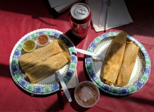 Two plates of tamales with sauces, accompanied by a can of soda, on a red tablecloth.