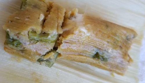Close-up of a tamale, showing a filling of green ingredients wrapped in corn masa and corn husk.