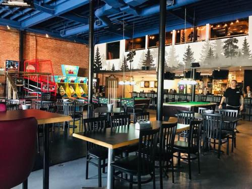 Interior of a casual restaurant with tables, arcade games, and a rustic decor featuring forest-themed murals.