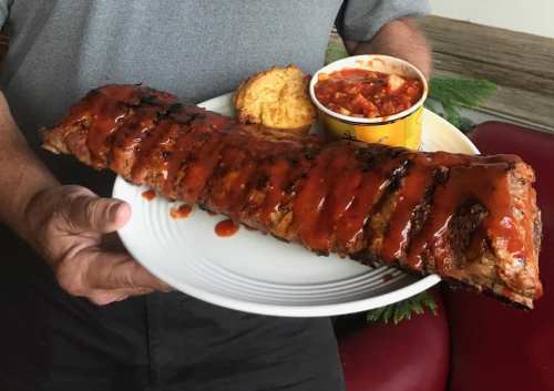 A person holds a large plate of ribs covered in barbecue sauce, accompanied by cornbread and a cup of baked beans.