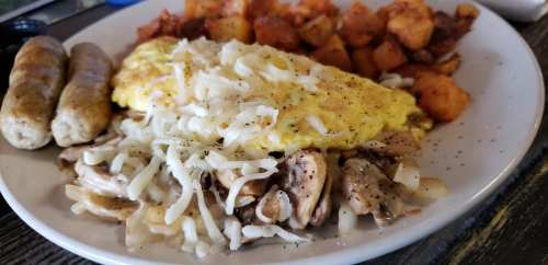 A plate of breakfast featuring an omelet with cheese, sautéed mushrooms, and two sausage links, alongside seasoned potatoes.