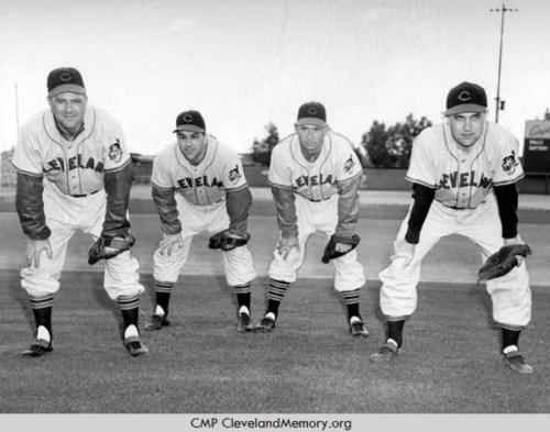 Four baseball players in uniforms pose on a field, crouching with gloves, ready for action.