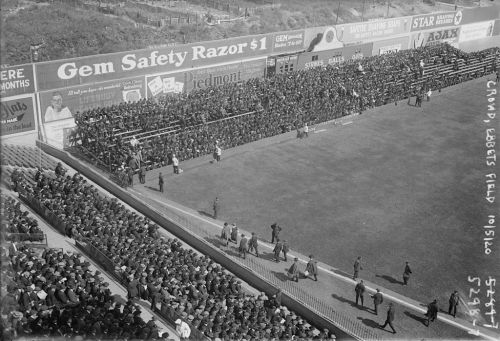 A large crowd gathers at a baseball field, with spectators seated in bleachers and standing along the sidelines.
