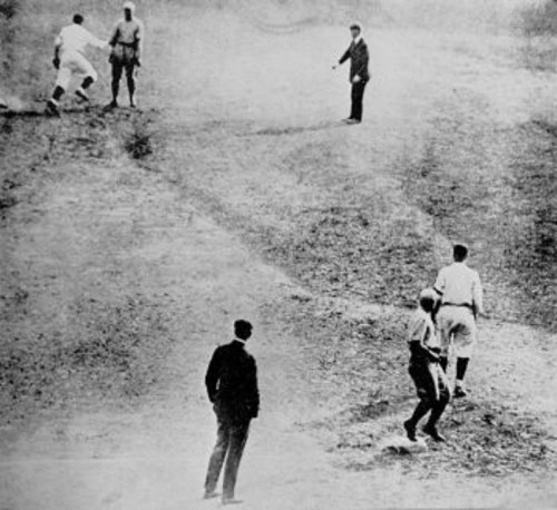 A historic black-and-white photo of a baseball game, showing players on the field and an umpire.
