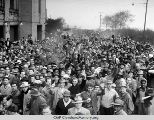 A large crowd of men in hats, gathered outdoors, some waving, in a historic black-and-white photograph.