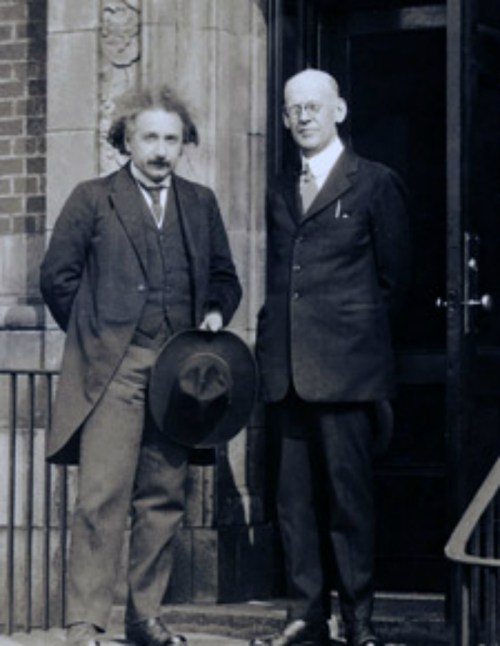 Two men in early 20th-century attire stand outside a building, one holding a hat, both smiling at the camera.