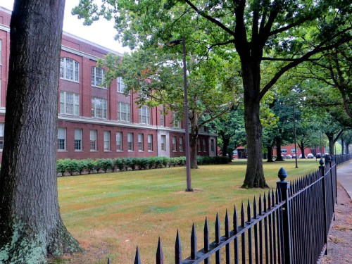 A brick school building surrounded by trees and a grassy area, with a black fence in the foreground.