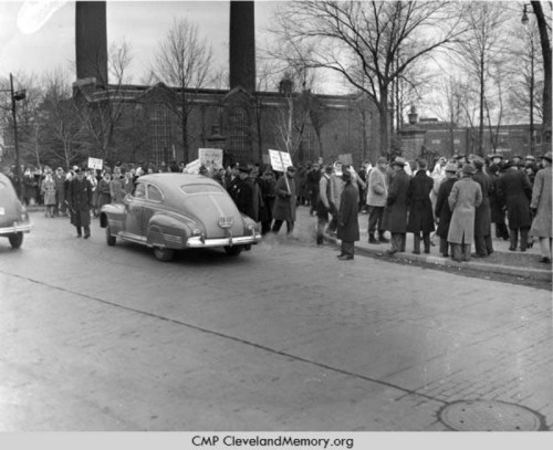 A vintage car drives past a crowd holding signs in front of industrial buildings, set in a winter landscape.