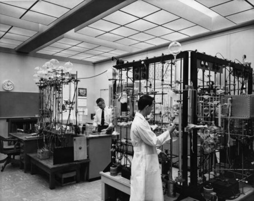 Two scientists in lab coats work in a vintage laboratory filled with glass equipment and scientific apparatus.