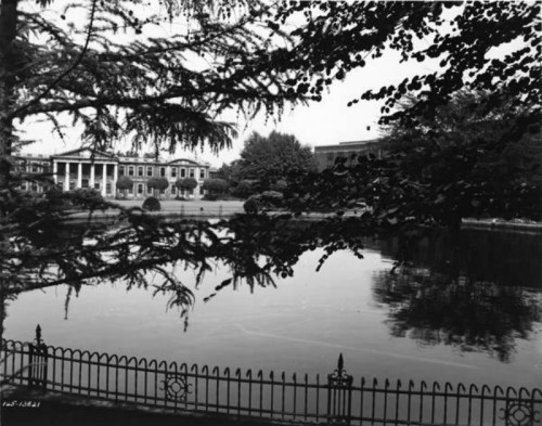 A serene view of a pond surrounded by trees, with a historic building in the background and a decorative fence in the foreground.