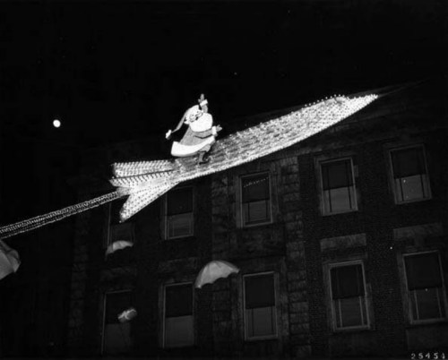 A vintage black-and-white photo of Santa on a rocket, illuminated with lights, above a building at night.
