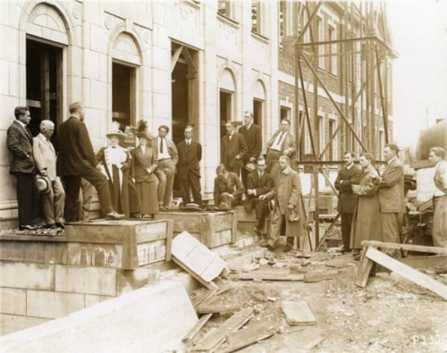 A historical black-and-white photo of a group of men gathered outside a building under construction, discussing.