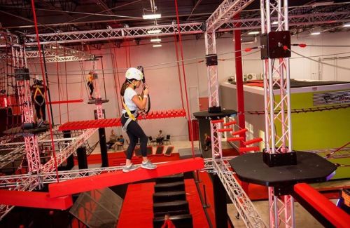 A person in a helmet navigates a high ropes course with red and black obstacles in an indoor adventure park.