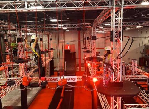 Two people navigate an indoor obstacle course with ropes and platforms, surrounded by red lights and metal structures.