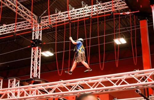 A person navigates a high ropes course, suspended on cables and beams in a brightly lit indoor facility.