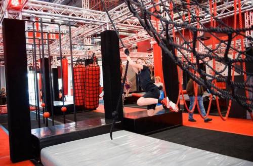 A person jumps on an obstacle course with ropes and nets in a vibrant indoor gym setting.