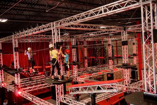 A group of people navigating a high ropes course with safety gear, surrounded by metal structures and red lighting.