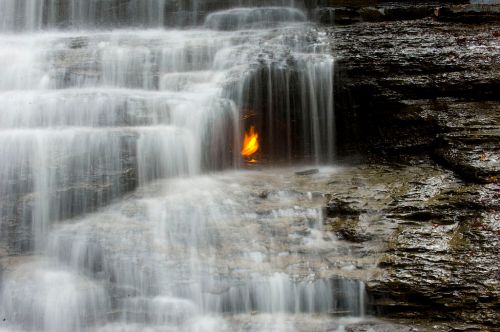 A waterfall cascades over rocks, with a small flame flickering amidst the flowing water.