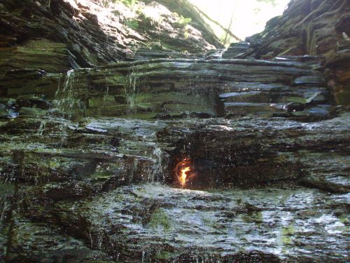 A rocky cliff with cascading water and a small flame visible among the stones, surrounded by greenery.