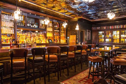 A stylish bar interior featuring a well-stocked shelf of spirits, leather stools, and warm lighting.