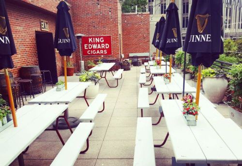 Outdoor patio with picnic tables, umbrellas, and potted plants, featuring a sign for King Edward Cigars.