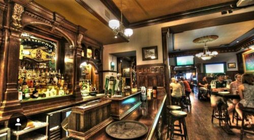 A cozy bar interior with wooden decor, a well-stocked bar, and patrons enjoying drinks at tables.
