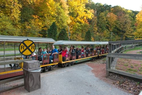 A colorful train filled with passengers travels through a scenic area with autumn foliage and a railroad crossing sign.
