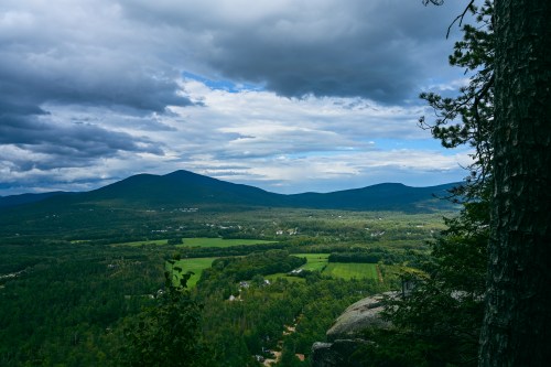 A panoramic view of green valleys and mountains under a cloudy sky, with trees in the foreground.