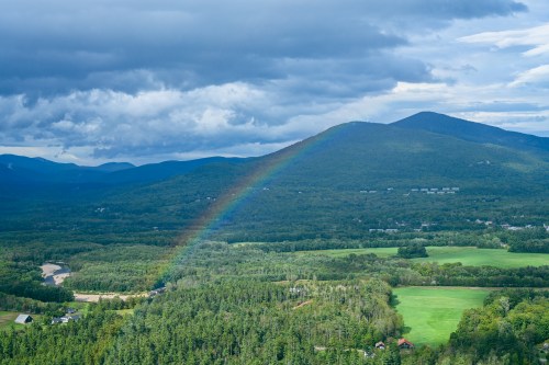 A vibrant rainbow arcs over lush green mountains and fields under a cloudy sky.