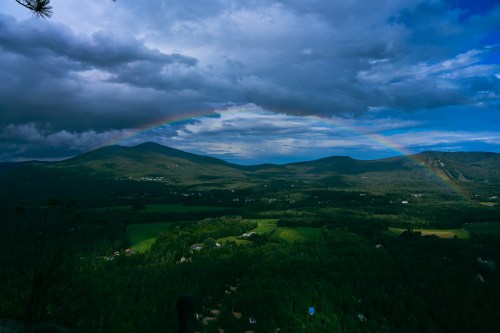 A vibrant rainbow arcs over green mountains and valleys under a cloudy sky.