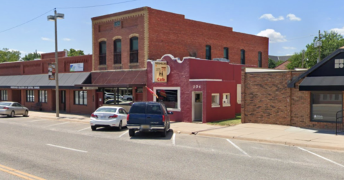 A street view of a red brick building with a café sign, surrounded by parked cars and other shops on a sunny day.