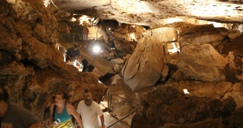 Visitors explore a rocky cave with illuminated pathways and large boulders, creating a natural underground landscape.