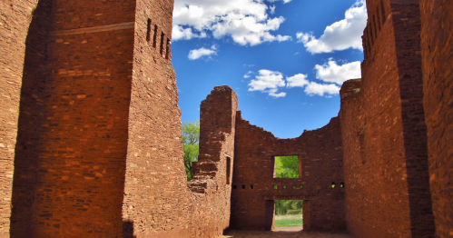 Ruins of a stone structure under a bright blue sky with scattered clouds, surrounded by greenery.