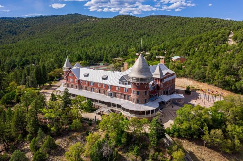 A large, historic building with turrets surrounded by lush green trees and mountains under a blue sky.