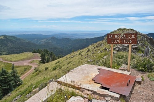 A scenic mountain view with a sign reading "La Mosca L.O. Elev. 11,036" overlooking a valley and distant hills.