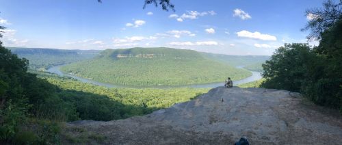 A panoramic view of a lush green valley with a river winding through it, and a person sitting on a rocky ledge.