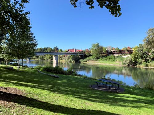 A serene riverside park with green grass, trees, a picnic table, and a bridge in the background under a clear blue sky.