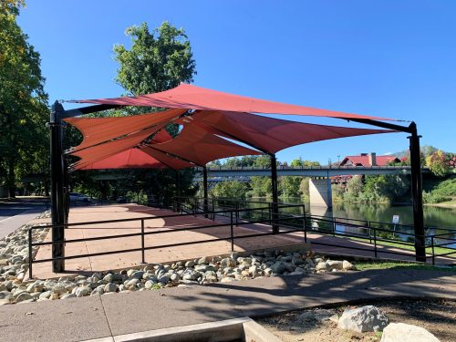 A shaded pavilion with red fabric canopies overlooking a river and bridge, surrounded by trees and a stone path.
