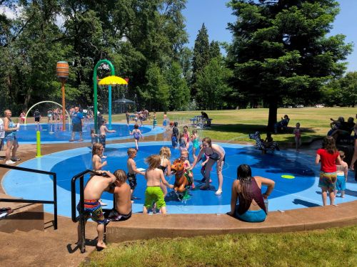 Children and adults enjoy a sunny day at a splash pad, playing in water and having fun in a park setting.