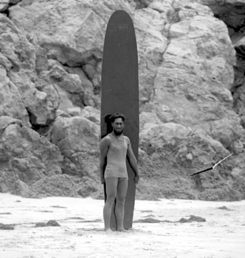 A man stands on the beach with a surfboard, surrounded by rocky cliffs in a black and white photograph.