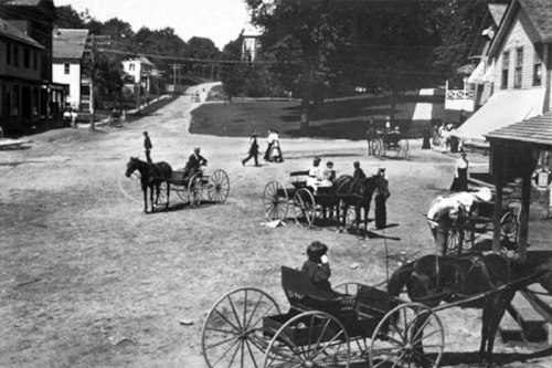 A historic black-and-white photo of a village square with horse-drawn carriages and people socializing.