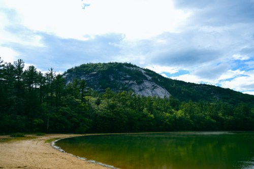 A serene lake surrounded by lush green trees and a rocky mountain under a cloudy sky.