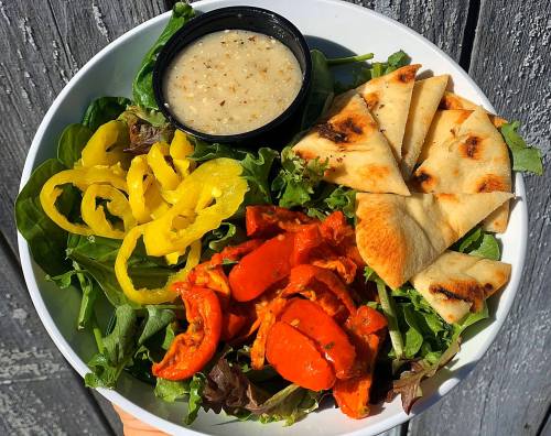 A bowl of mixed greens topped with roasted red peppers, yellow peppers, pita bread, and a small container of dressing.
