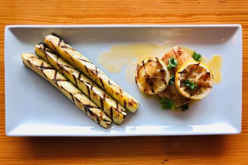 Grilled asparagus and lemon slices served with a piece of fish on a white plate, set on a wooden table.