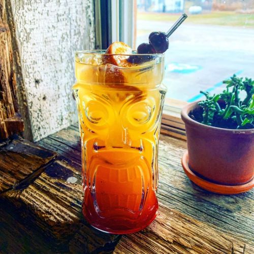 A colorful tiki cocktail in a decorative glass, garnished with cherries and orange slices, next to a small potted plant.