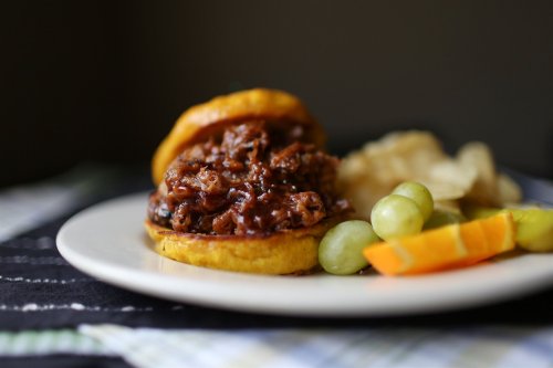A pulled pork sandwich on a bun, served with grapes, orange slices, pickles, and potato chips on a white plate.