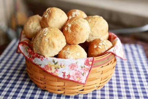 A woven basket filled with golden-brown bread rolls, topped with oats, resting on a checkered tablecloth.