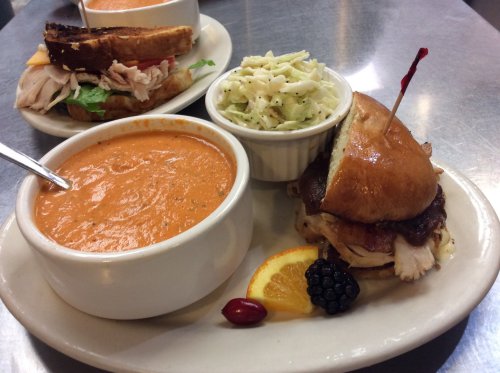 A plate with a sandwich, coleslaw, and a bowl of tomato soup, garnished with fruit.