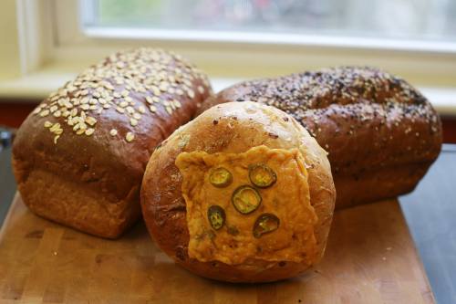Three loaves of bread on a wooden cutting board: one topped with oats, one with seeds, and one with cheese and jalapeños.